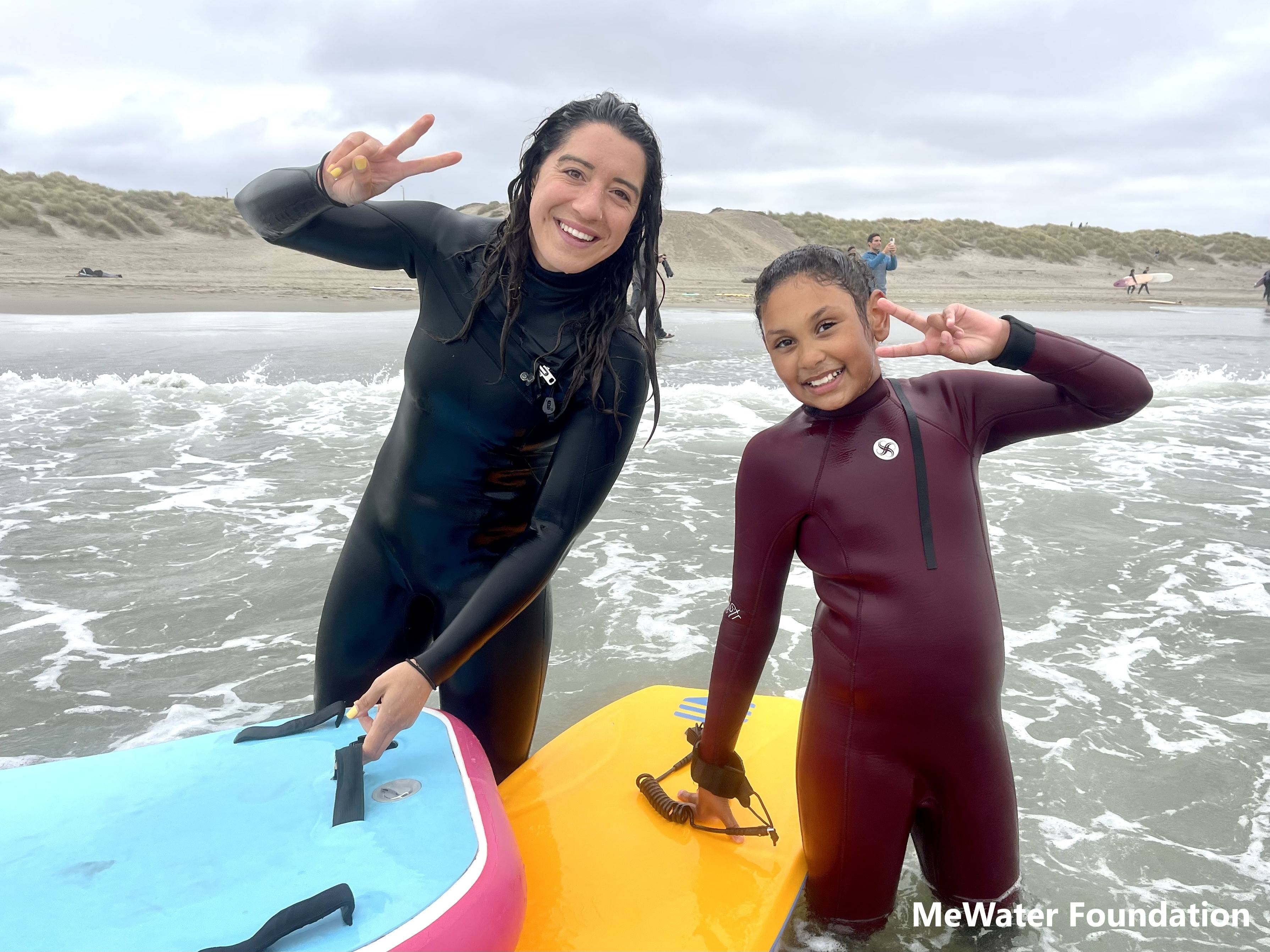 A young person and an adult stand in the surf wearing wetsuits and holding boogie boards while smiling for the camera. Photo: MeWater Foundation.